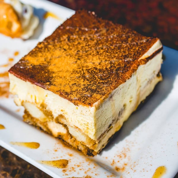 A rectangular slice of tiramisu, dusted with cocoa powder, on a white plate with caramel drizzle.