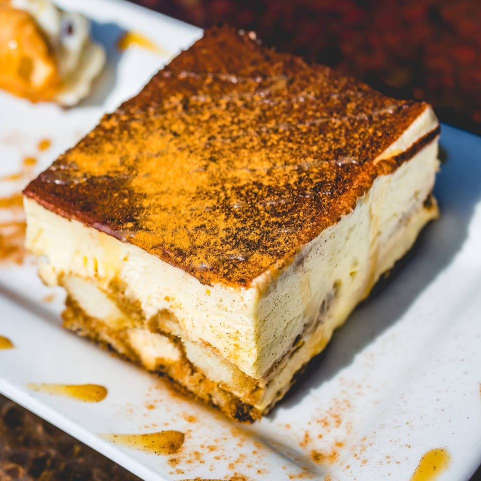 A rectangular slice of tiramisu, dusted with cocoa powder, on a white plate with caramel drizzle.